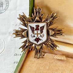 Vintage gold‑tone heraldic brooch with white enamel shield and red‑gold griffin coat of arms, photographed on antique paper and boxes.