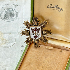 Vintage gold‑tone heraldic brooch with white enamel shield and red‑gold griffin coat of arms, photographed on antique paper and boxes.