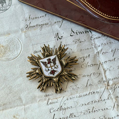 Vintage gold‑tone heraldic brooch with white enamel shield and red‑gold griffin coat of arms, photographed on antique paper and boxes.