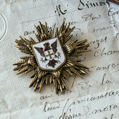 Vintage gold‑tone heraldic brooch with white enamel shield and red‑gold griffin coat of arms, photographed on antique paper and boxes.