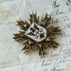 Vintage gold‑tone heraldic brooch with white enamel shield and red‑gold griffin coat of arms, photographed on antique paper and boxes.