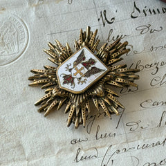Vintage gold‑tone heraldic brooch with white enamel shield and red‑gold griffin coat of arms, photographed on antique paper and boxes.