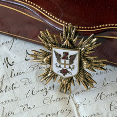 Vintage gold‑tone heraldic brooch with white enamel shield and red‑gold griffin coat of arms, photographed on antique paper and boxes.