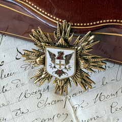 Vintage gold‑tone heraldic brooch with white enamel shield and red‑gold griffin coat of arms, photographed on antique paper and boxes.
