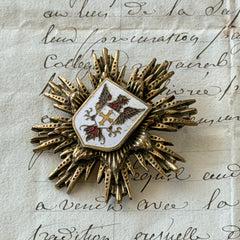 Vintage gold‑tone heraldic brooch with white enamel shield and red‑gold griffin coat of arms, photographed on antique paper and boxes.