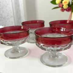 Set of vintage Indiana Glass cranberry sherbet glasses with thumbprint detailing, styled with soft lighting and floral accents to highlight their color, texture, and collectible charm.