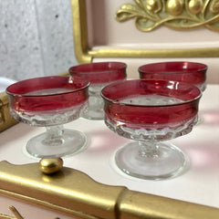 Set of vintage Indiana Glass cranberry sherbet glasses with thumbprint detailing, styled with soft lighting and floral accents to highlight their color, texture, and collectible charm.
