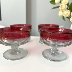 Set of vintage Indiana Glass cranberry sherbet glasses with thumbprint detailing, styled with soft lighting and floral accents to highlight their color, texture, and collectible charm.