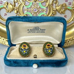 Vintage oval Italian micro‑mosaic clip‑on earrings with yellow and blue floral designs on a green background, framed in a gold‑tone twisted rope border. The mosaic is intact and detailed, with light wear visible on the backs.