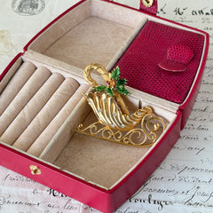 Vintage JJ Jonette gold‑tone sleigh brooch with green enamel holly leaves, red rhinestone berries, and clear rhinestones, photographed on antique script and red box background.