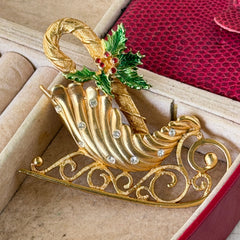 Vintage JJ Jonette gold‑tone sleigh brooch with green enamel holly leaves, red rhinestone berries, and clear rhinestones, photographed on antique script and red box background.