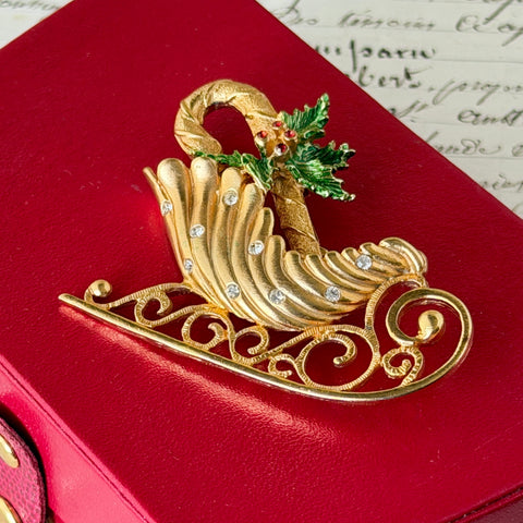 Vintage JJ Jonette gold‑tone sleigh brooch with green enamel holly leaves, red rhinestone berries, and clear rhinestones, photographed on antique script and red box background.