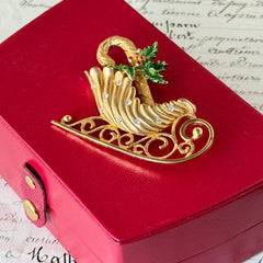Vintage JJ Jonette gold‑tone sleigh brooch with green enamel holly leaves, red rhinestone berries, and clear rhinestones, photographed on antique script and red box background.