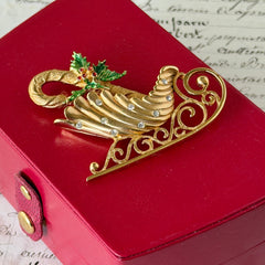 Vintage JJ Jonette gold‑tone sleigh brooch with green enamel holly leaves, red rhinestone berries, and clear rhinestones, photographed on antique script and red box background.