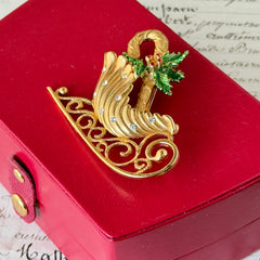 Vintage JJ Jonette gold‑tone sleigh brooch with green enamel holly leaves, red rhinestone berries, and clear rhinestones, photographed on antique script and red box background.