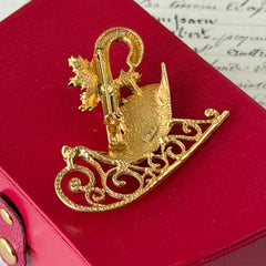 Vintage JJ Jonette gold‑tone sleigh brooch with green enamel holly leaves, red rhinestone berries, and clear rhinestones, photographed on antique script and red box background.