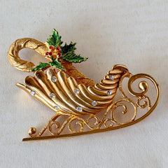 Vintage JJ Jonette gold‑tone sleigh brooch with green enamel holly leaves, red rhinestone berries, and clear rhinestones, photographed on antique script and red box background.