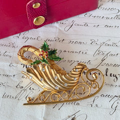 Vintage JJ Jonette gold‑tone sleigh brooch with green enamel holly leaves, red rhinestone berries, and clear rhinestones, photographed on antique script and red box background.