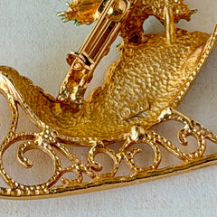 Vintage JJ Jonette gold‑tone sleigh brooch with green enamel holly leaves, red rhinestone berries, and clear rhinestones, photographed on antique script and red box background.
