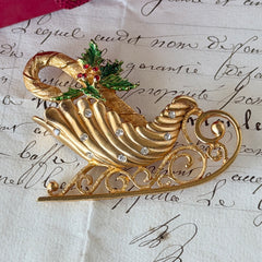 Vintage JJ Jonette gold‑tone sleigh brooch with green enamel holly leaves, red rhinestone berries, and clear rhinestones, photographed on antique script and red box background.