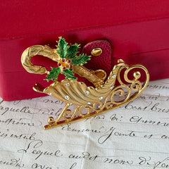 Vintage JJ Jonette gold‑tone sleigh brooch with green enamel holly leaves, red rhinestone berries, and clear rhinestones, photographed on antique script and red box background.