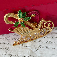 Vintage JJ Jonette gold‑tone sleigh brooch with green enamel holly leaves, red rhinestone berries, and clear rhinestones, photographed on antique script and red box background.