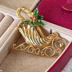 Vintage JJ Jonette gold‑tone sleigh brooch with green enamel holly leaves, red rhinestone berries, and clear rhinestones, photographed on antique script and red box background.