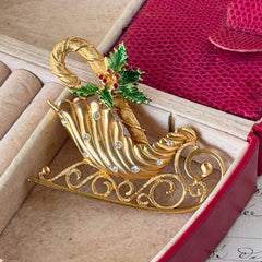 Vintage JJ Jonette gold‑tone sleigh brooch with green enamel holly leaves, red rhinestone berries, and clear rhinestones, photographed on antique script and red box background.