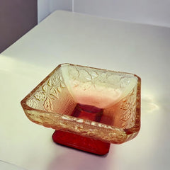 Vintage Jeannette Amberina Depression glass dish from the 1930s featuring a square footed base, an Acorn and Leaf pattern, and a color gradient from amber/yellow to red. Perfect as a candy dish or decorative piece, with a small chip on the rim adding character to its vintage charm.