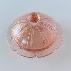 Vintage Jeannette Cherry Blossom pink Depression glass butter dish with embossed floral pattern, scalloped edges, and domed lid, shown in natural light on a white tabletop.