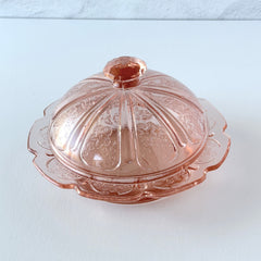 Vintage Jeannette Cherry Blossom pink Depression glass butter dish with embossed floral pattern, scalloped edges, and domed lid, shown in natural light on a white tabletop.