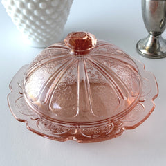 Vintage Jeannette Cherry Blossom pink Depression glass butter dish with embossed floral pattern, scalloped edges, and domed lid, shown in natural light on a white tabletop.