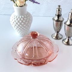 Vintage Jeannette Cherry Blossom pink Depression glass butter dish with embossed floral pattern, scalloped edges, and domed lid, shown in natural light on a white tabletop.