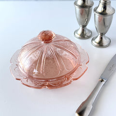 Vintage Jeannette Cherry Blossom pink Depression glass butter dish with embossed floral pattern, scalloped edges, and domed lid, shown in natural light on a white tabletop.