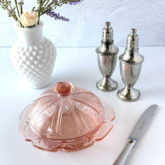 Vintage Jeannette Cherry Blossom pink Depression glass butter dish with embossed floral pattern, scalloped edges, and domed lid, shown in natural light on a white tabletop.