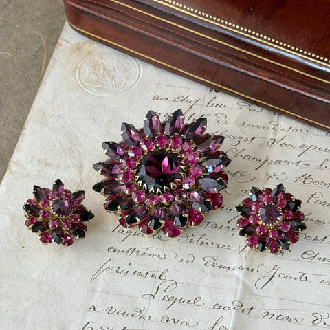 Vintage Judy Lee demi parure featuring a gold‑tone floral brooch with layered pink and amethyst rhinestones, paired with matching clip‑on earrings. The set sparkles in natural light, showing vivid color, faceted shine, and mid‑century detailing.
