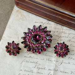 Vintage Judy Lee demi parure featuring a gold‑tone floral brooch with layered pink and amethyst rhinestones, paired with matching clip‑on earrings. The set sparkles in natural light, showing vivid color, faceted shine, and mid‑century detailing.
