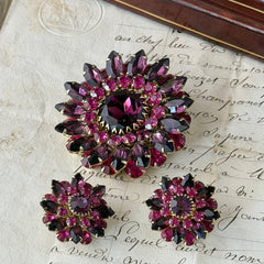 Vintage Judy Lee demi parure featuring a gold‑tone floral brooch with layered pink and amethyst rhinestones, paired with matching clip‑on earrings. The set sparkles in natural light, showing vivid color, faceted shine, and mid‑century detailing.