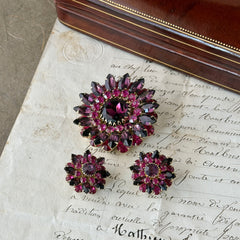 Vintage Judy Lee demi parure featuring a gold‑tone floral brooch with layered pink and amethyst rhinestones, paired with matching clip‑on earrings. The set sparkles in natural light, showing vivid color, faceted shine, and mid‑century detailing.