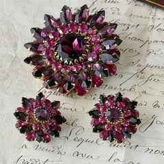 Vintage Judy Lee demi parure featuring a gold‑tone floral brooch with layered pink and amethyst rhinestones, paired with matching clip‑on earrings. The set sparkles in natural light, showing vivid color, faceted shine, and mid‑century detailing.