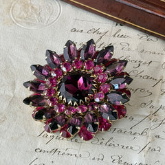 Vintage Judy Lee demi parure featuring a gold‑tone floral brooch with layered pink and amethyst rhinestones, paired with matching clip‑on earrings. The set sparkles in natural light, showing vivid color, faceted shine, and mid‑century detailing.
