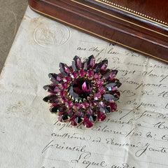 Vintage Judy Lee demi parure featuring a gold‑tone floral brooch with layered pink and amethyst rhinestones, paired with matching clip‑on earrings. The set sparkles in natural light, showing vivid color, faceted shine, and mid‑century detailing.