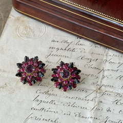 Vintage Judy Lee demi parure featuring a gold‑tone floral brooch with layered pink and amethyst rhinestones, paired with matching clip‑on earrings. The set sparkles in natural light, showing vivid color, faceted shine, and mid‑century detailing.
