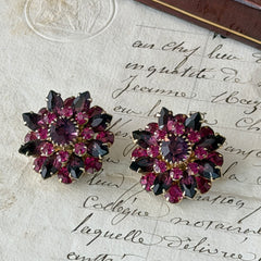 Vintage Judy Lee demi parure featuring a gold‑tone floral brooch with layered pink and amethyst rhinestones, paired with matching clip‑on earrings. The set sparkles in natural light, showing vivid color, faceted shine, and mid‑century detailing.