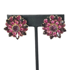 Vintage Judy Lee demi parure featuring a gold‑tone floral brooch with layered pink and amethyst rhinestones, paired with matching clip‑on earrings. The set sparkles in natural light, showing vivid color, faceted shine, and mid‑century detailing.
