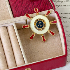 Vintage Louis Giusti gold‑tone ship‑wheel brooch with white, red, and navy enamel and anchor motif, displayed on antique paper background.