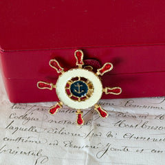 Vintage Louis Giusti gold‑tone ship‑wheel brooch with white, red, and navy enamel and anchor motif, displayed on antique paper background.