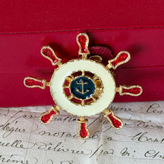 Vintage Louis Giusti gold‑tone ship‑wheel brooch with white, red, and navy enamel and anchor motif, displayed on antique paper background.