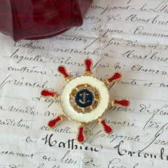 Vintage Louis Giusti gold‑tone ship‑wheel brooch with white, red, and navy enamel and anchor motif, displayed on antique paper background.
