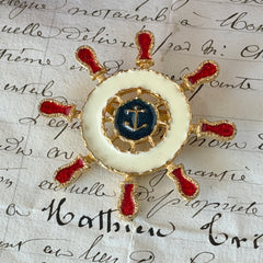 Vintage Louis Giusti gold‑tone ship‑wheel brooch with white, red, and navy enamel and anchor motif, displayed on antique paper background.